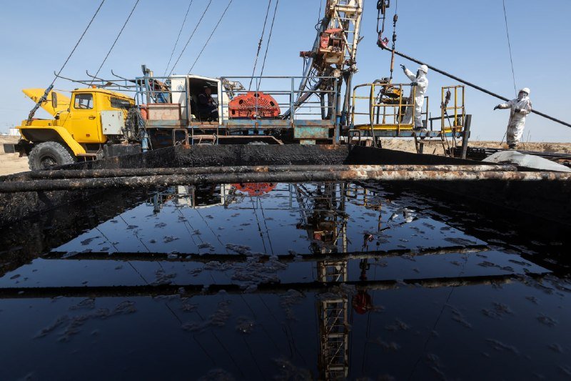Workers in white coveralls operate drilling equipment beside a dark, reflective liquid pool at an industrial site under a clear blue sky.
