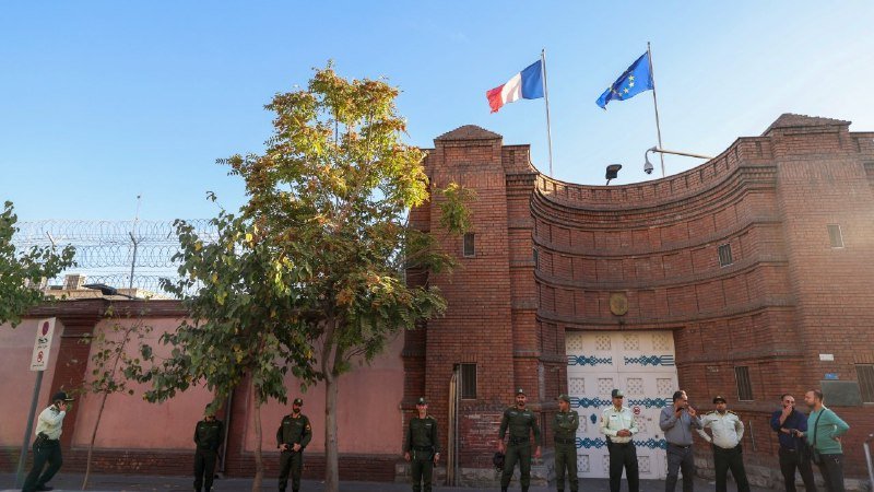 Uniformed guards and civilians stand outside a brick building topped with French and EU flags under clear blue skies.