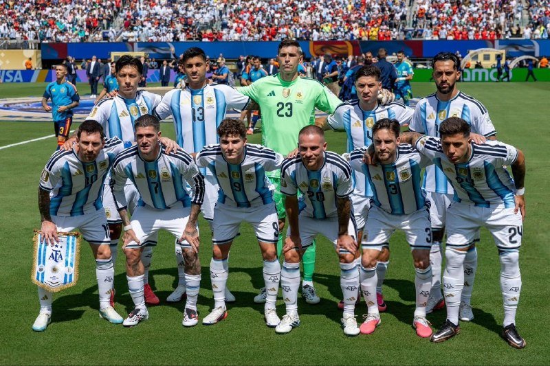 The Argentina national soccer team poses together on a soccer field before a match, wearing their blue and white striped jerseys with the AFA crest.