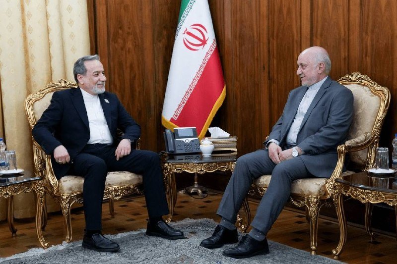 Two men in suits sit facing each other in ornate armchairs in front of an Iranian flag, with a telephone and teapot on a table between them.
