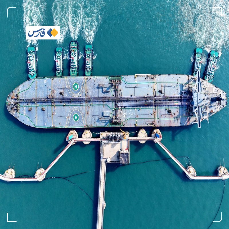 Aerial view of a large oil tanker docked at an offshore marine terminal, with tugboats maneuvering alongside in turquoise water; a Persian-language news logo appears in the upper corner.
