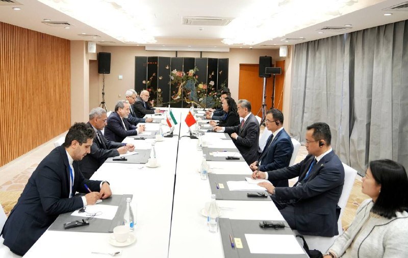 Two delegations in business attire sit across a long conference table in a meeting room, with small Iranian and Indonesian flags displayed at the center.
