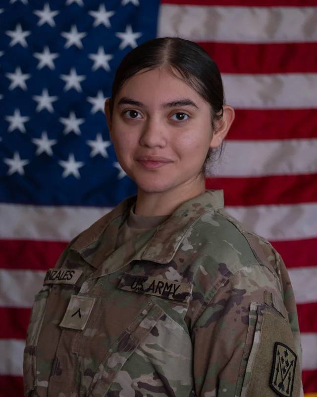 A woman in a camouflage U.S. Army uniform with visible name and unit patches stands posed in front of an American flag.