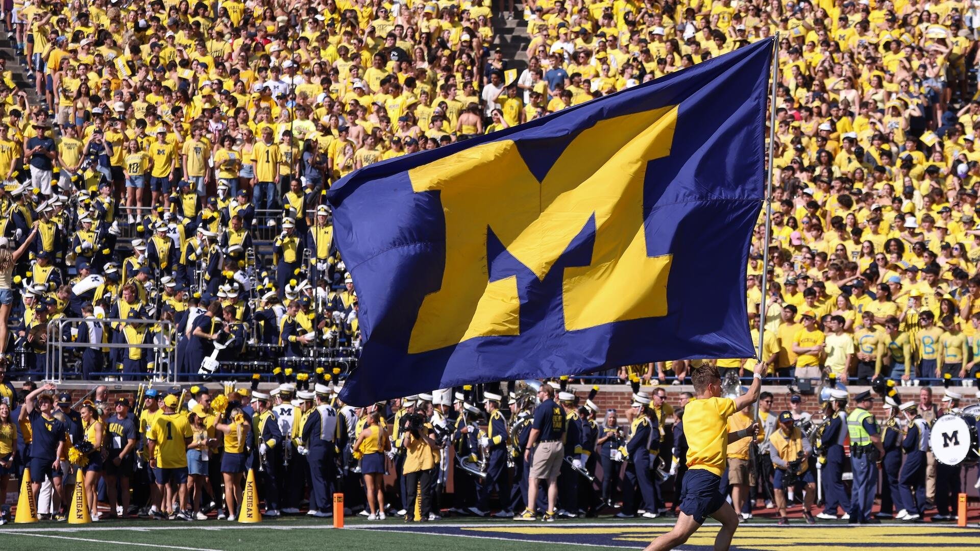 A person waves a large blue and yellow "M" flag before a marching band and a stadium crowd wearing matching yellow shirts.