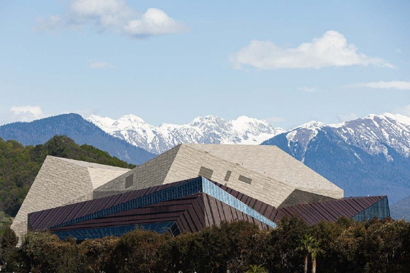 A modern building with angular, geometric rooftops and a textured stone-and-glass facade stands before a backdrop of snow-capped mountains under a partly cloudy sky.