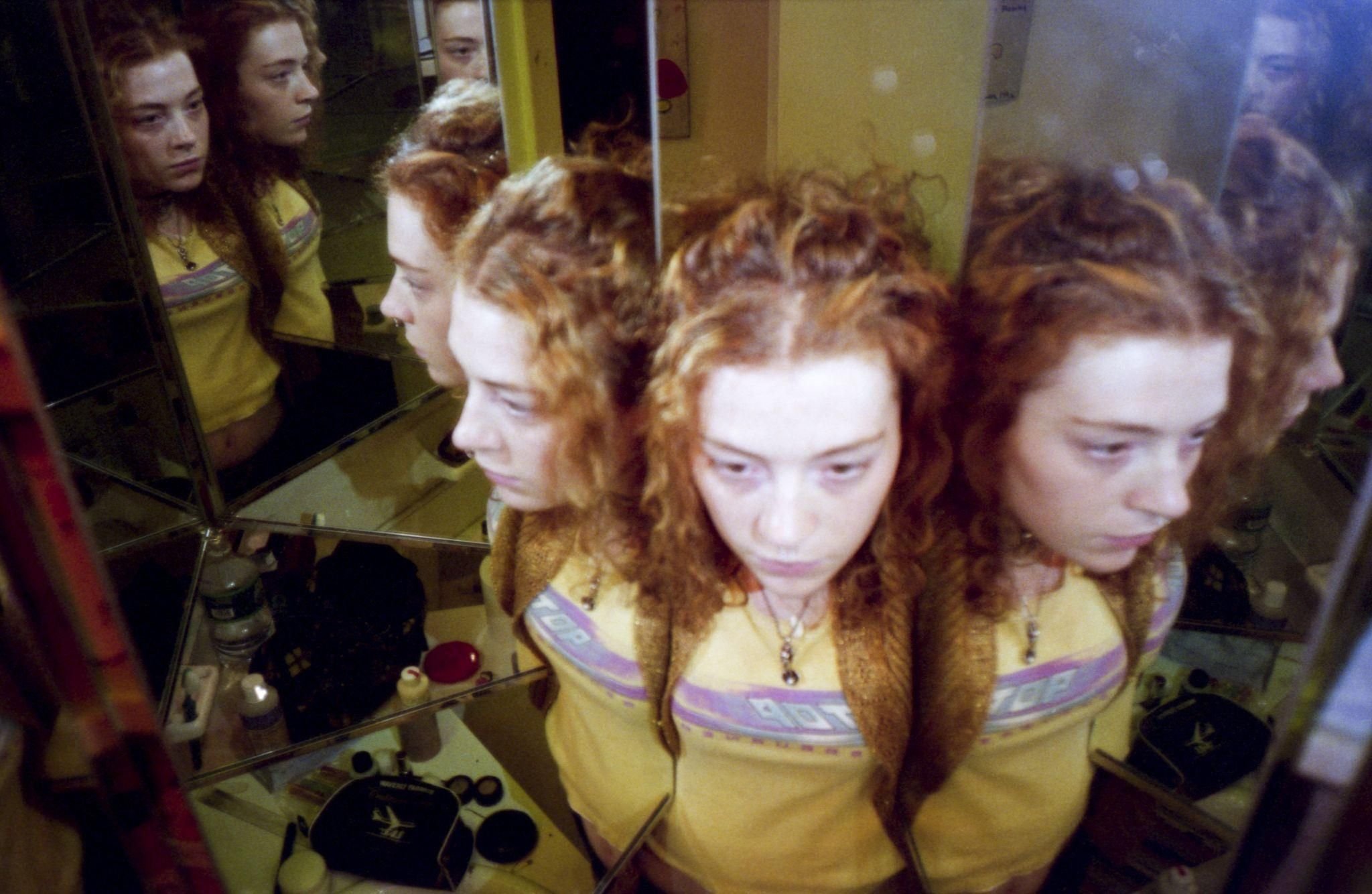 A young woman with curly auburn hair and a yellow graphic t-shirt is reflected multiple times in a mirrored wall, standing in a bathroom with shelves of toiletries.