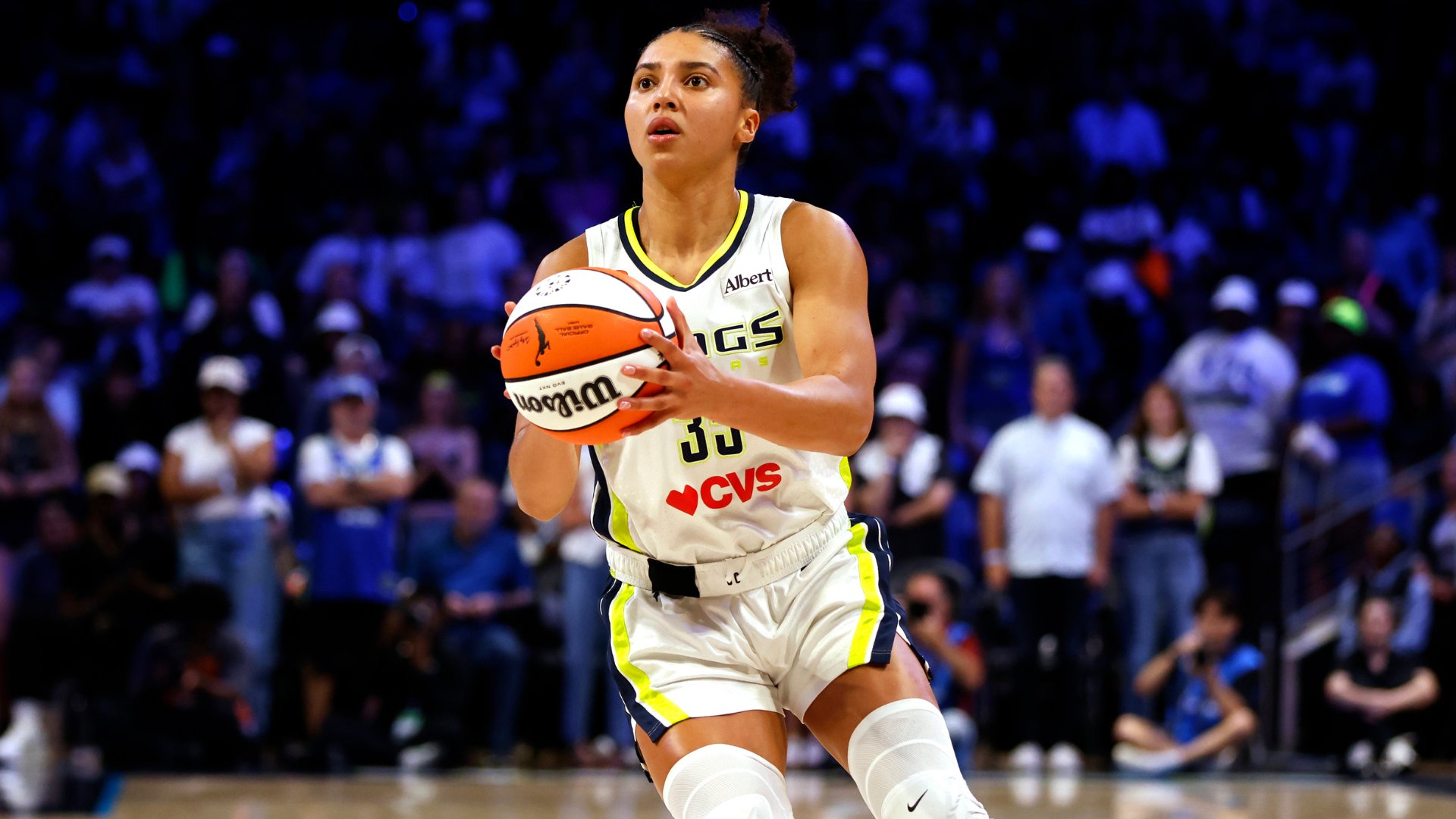 A female basketball player in a white Wings jersey prepares to shoot a basketball during a WNBA game, with a blurred crowd visible in the background.