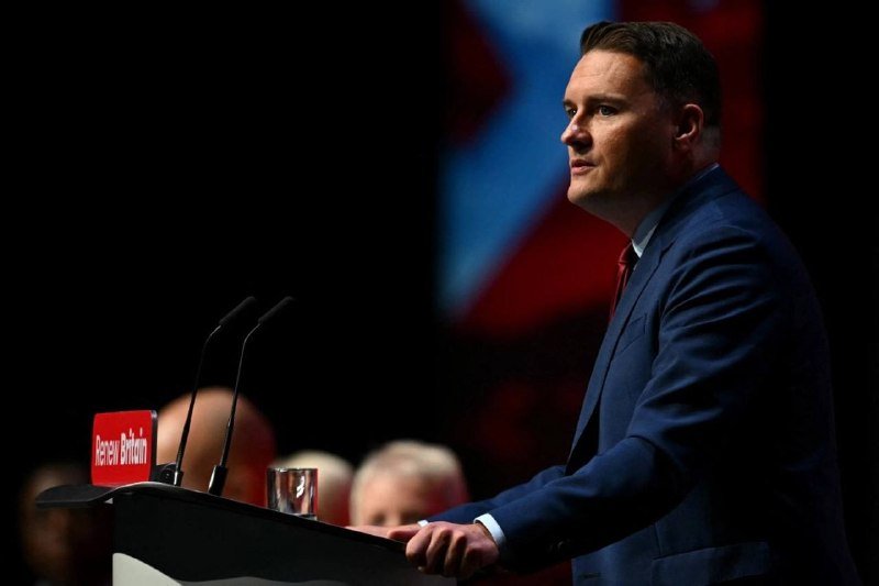 A man in a blue suit speaks at a podium displaying a "Renew Britain" placard, with a blurred flag visible behind him.