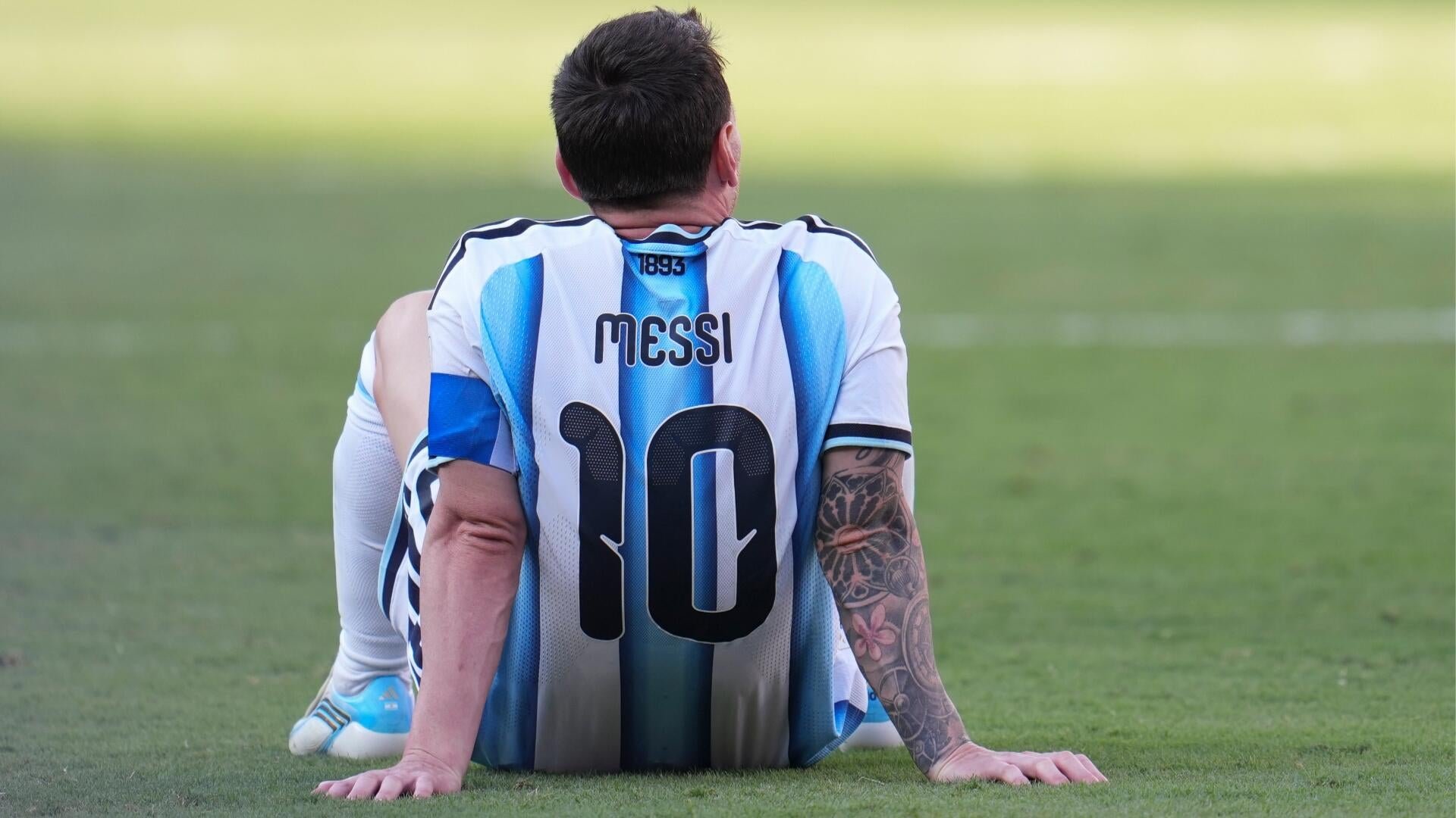 A soccer player wearing a light blue and white striped jersey with "MESSI" and the number 10 sits on a grass field, viewed from behind, with a tattooed arm visible.