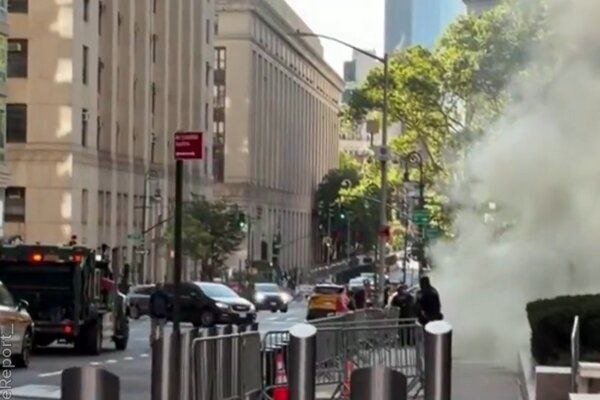 A large plume of white steam rises on a city street lined with tall buildings, traffic lights, vehicles, and pedestrians behind metal barricades.