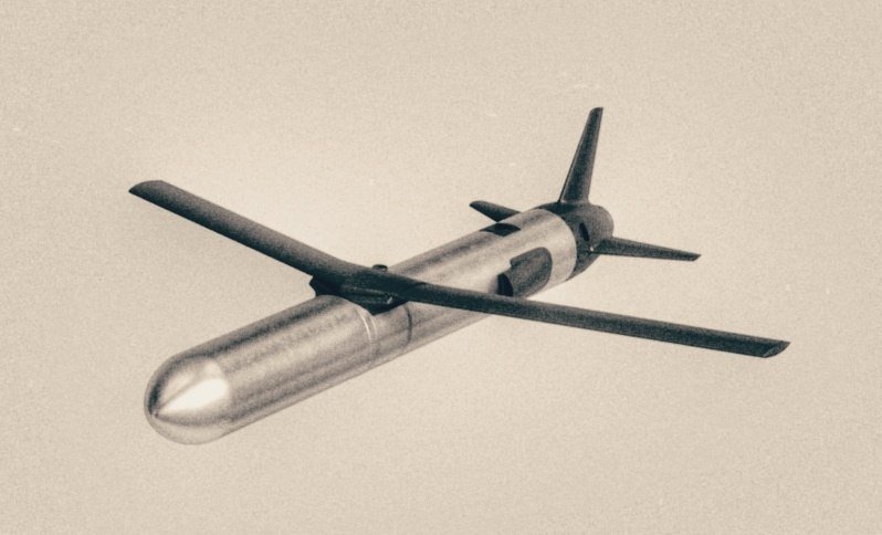 A missile with a silver cylindrical body, dark cruciform wings, and tail fins is shown in flight against a pale sky in this black-and-white photograph.