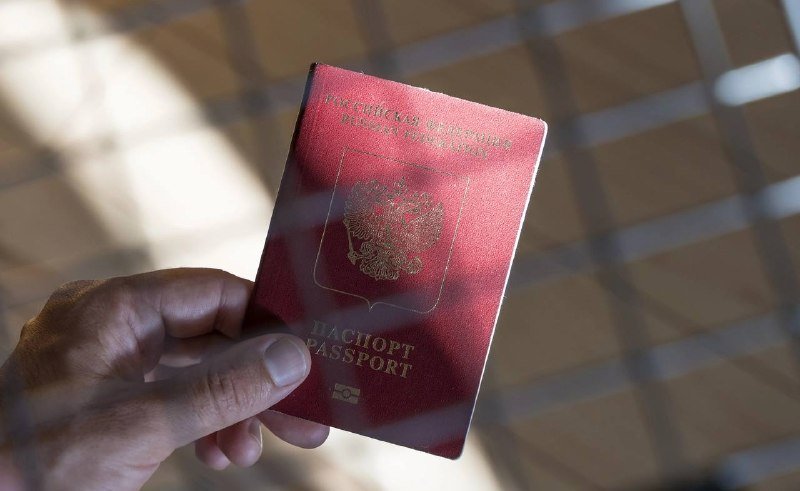 A hand holds a burgundy Russian passport with the gold double-headed eagle emblem, photographed against a blurred tiled floor.