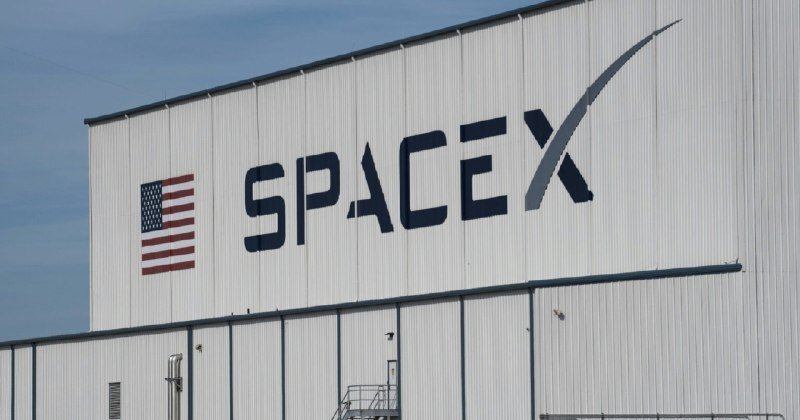The word "SPACEX" and an American flag are displayed on the side of a large white industrial building under a blue sky.