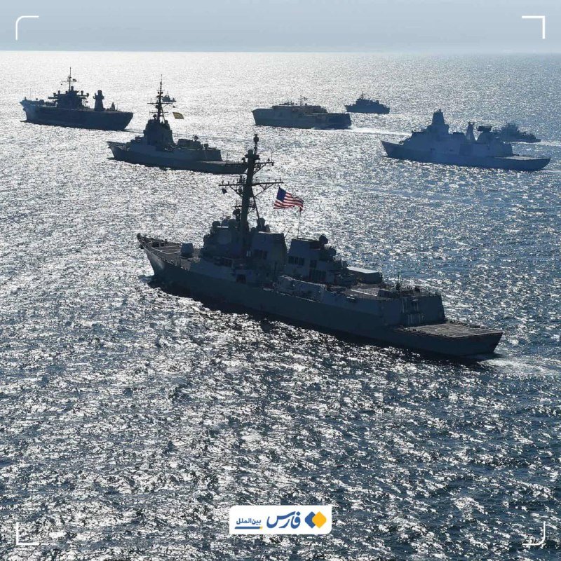 A formation of seven naval warships sails in a line across sunlit open ocean waters, with a U.S. flag visible on the nearest vessel's mast.