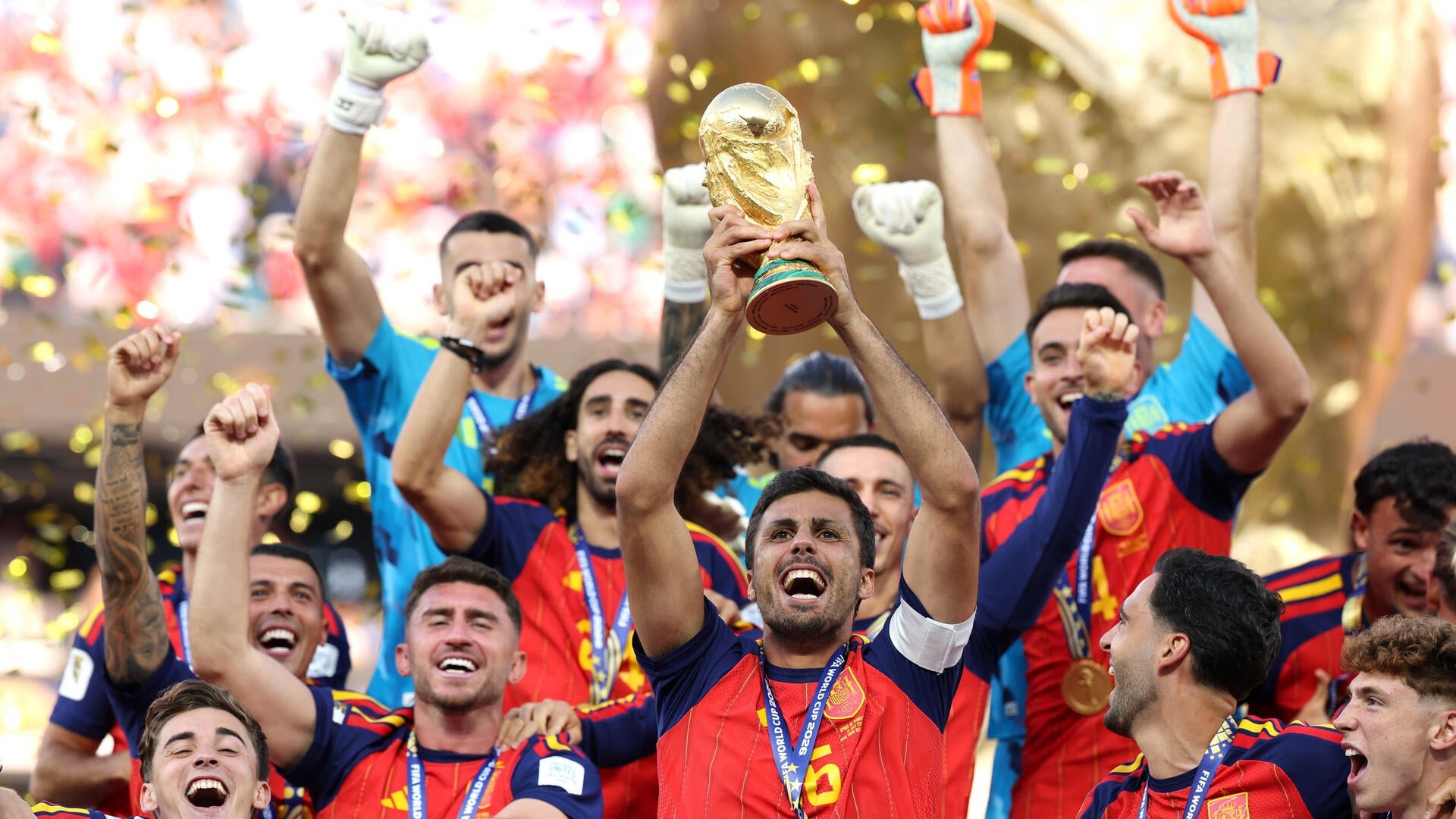 A team in red jerseys celebrates around a player lifting the golden FIFA World Cup trophy amid falling confetti.