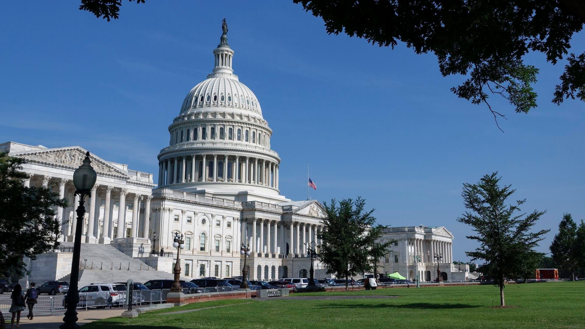 The U.S. Capitol building's white dome and columned facade stand under a clear blue sky, with an American flag visible and vehicles parked along the front.