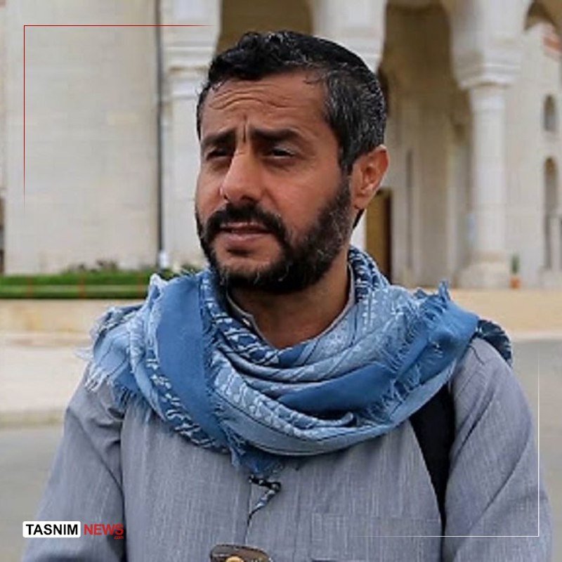 A bearded man wearing a blue scarf and gray shirt stands outdoors in front of a light-colored building with arched columns, with a "Tasnim News" logo visible.