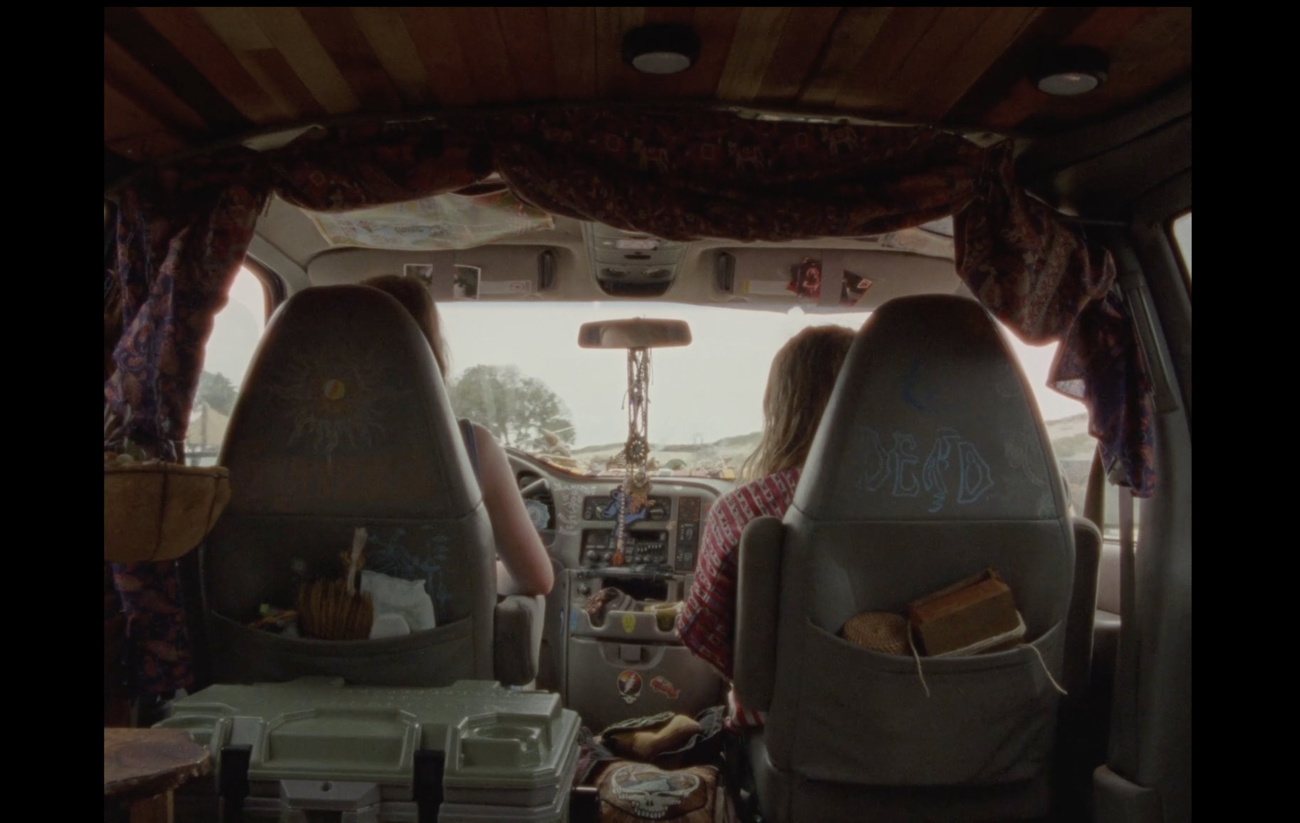 Two people sit in the front seats of a converted van with a wood-paneled ceiling, draped fabric, and storage cases visible in the rear.