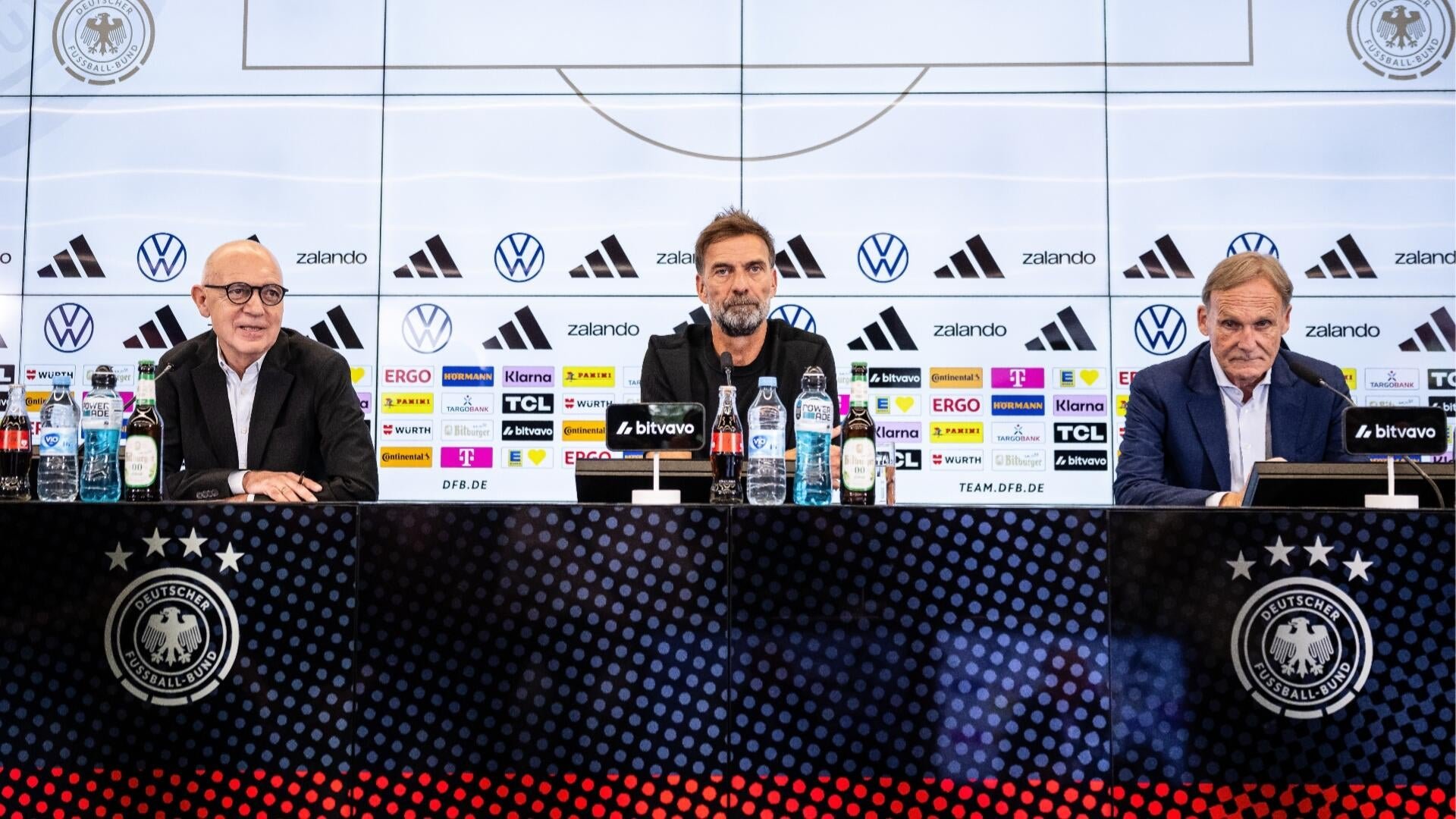 Three men sit at a press conference table branded with the Deutscher Fußball-Bund logo, accompanied by sponsor logos and microphones.