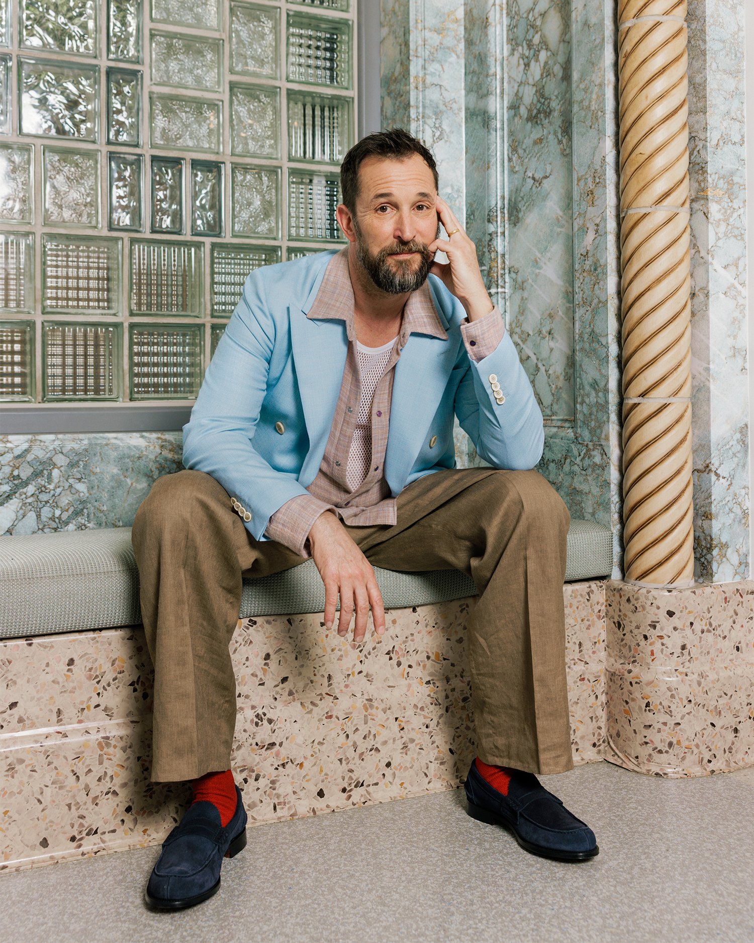 A bearded man in a light blue blazer, brown trousers, and red socks sits on a terrazzo ledge beside a glass block wall and twisted gold column.
