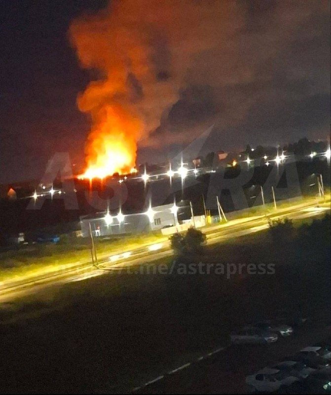 A large fire with bright orange flames and thick smoke rises above buildings at night, illuminating a nearby road lined with streetlights in a small town.