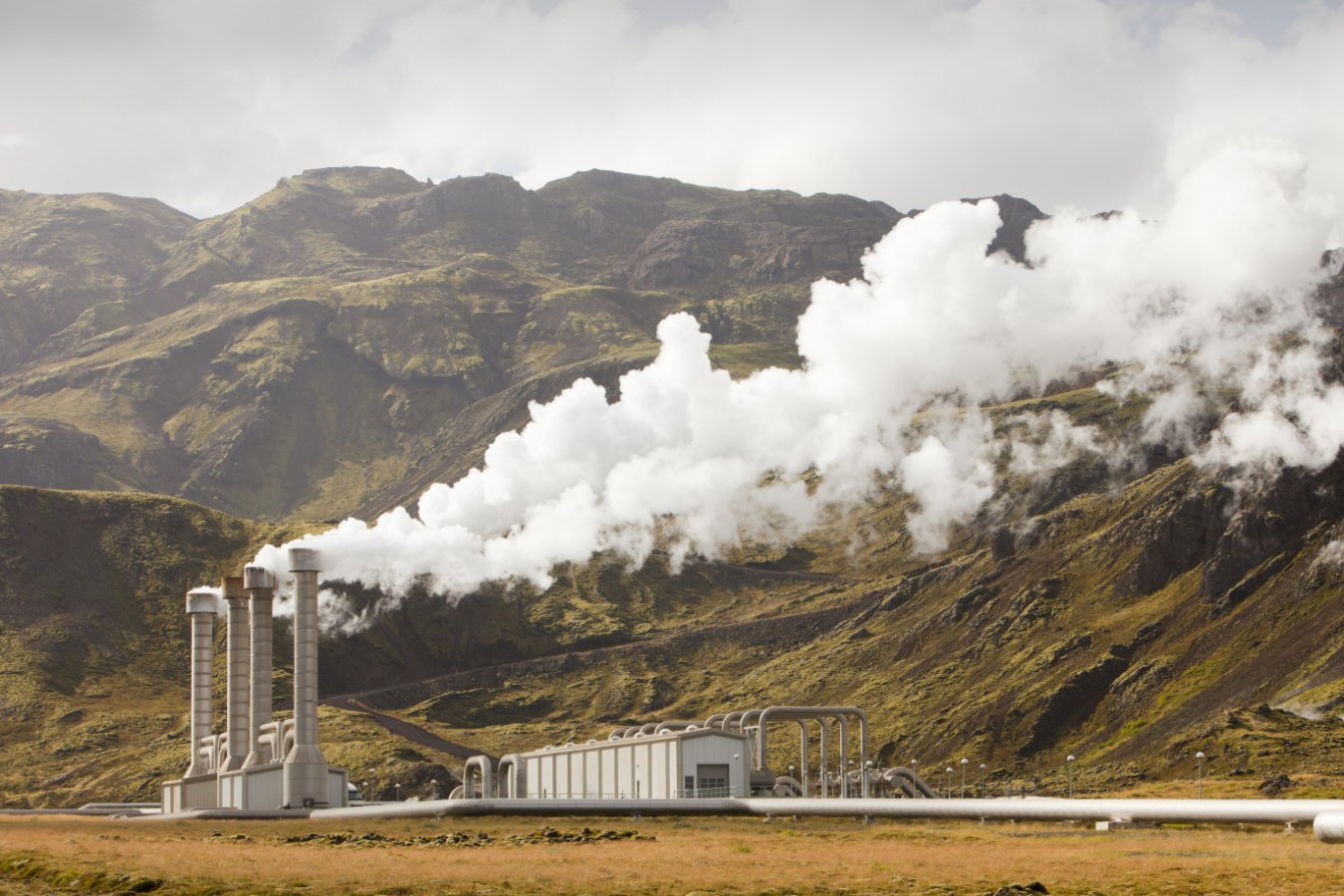 A geothermal power plant with tall smokestacks releases thick white steam into the sky, set against a backdrop of steep, moss-covered mountains under a cloudy sky.