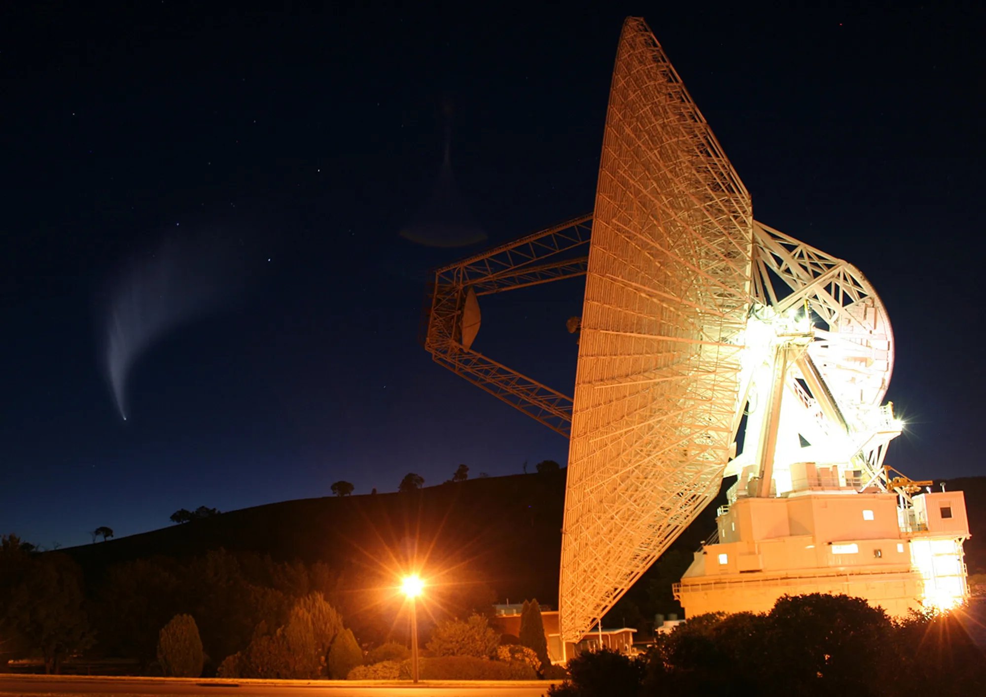 A large illuminated radio telescope dish stands at night with a bright comet visible in the starry sky above a darkened hillside.