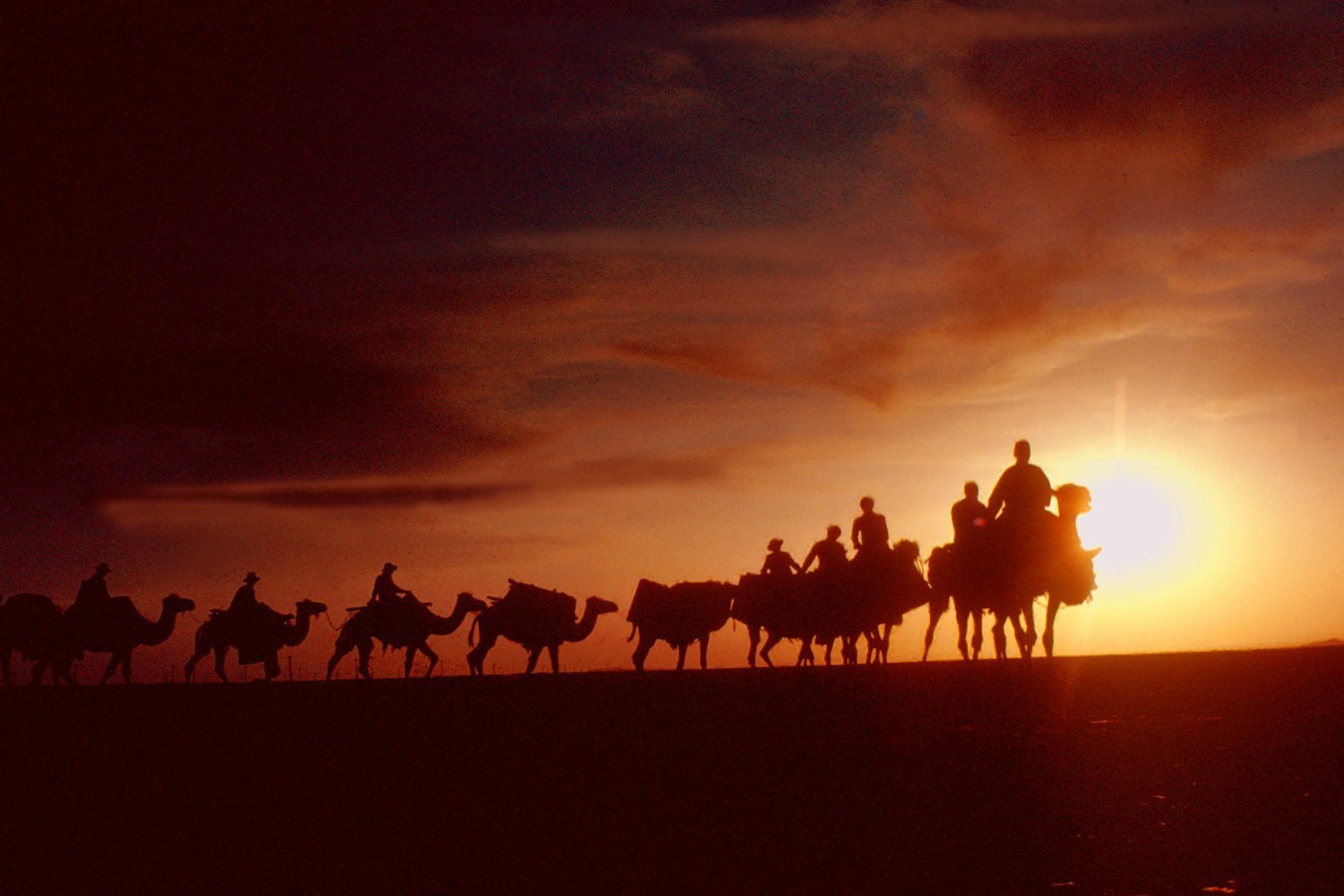 Silhouetted figures ride a caravan of camels across a ridgeline against a vivid orange sunset.
