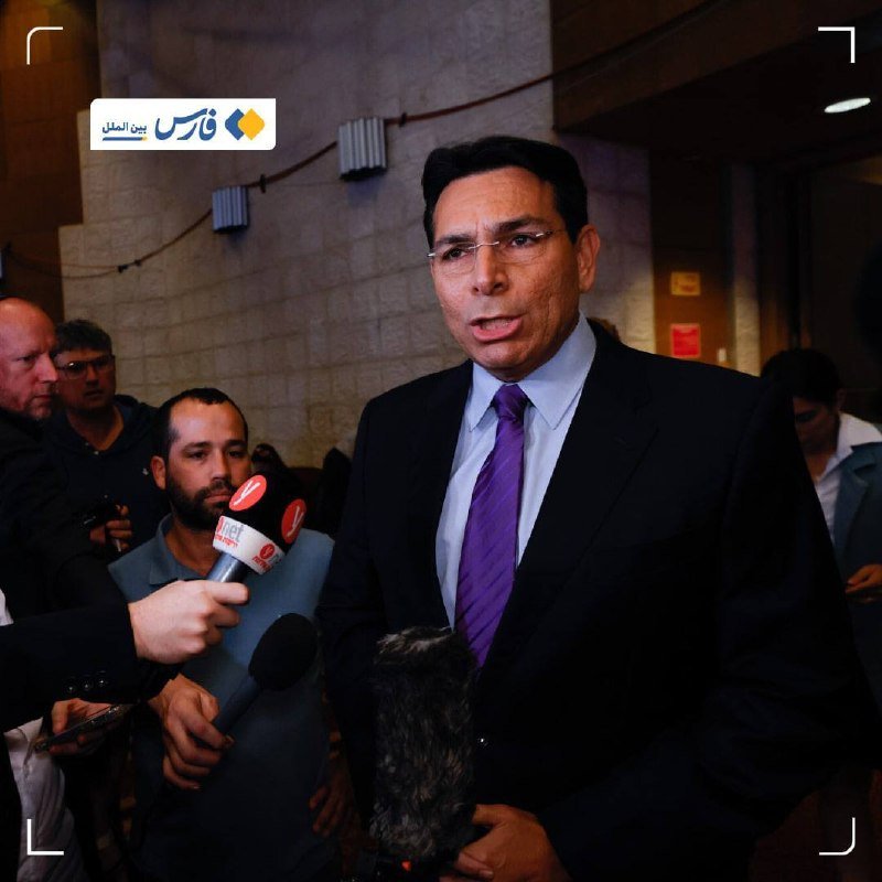 A bespectacled man in a dark suit and purple tie speaks to reporters holding microphones in a stone-walled hallway, accompanied by other individuals.