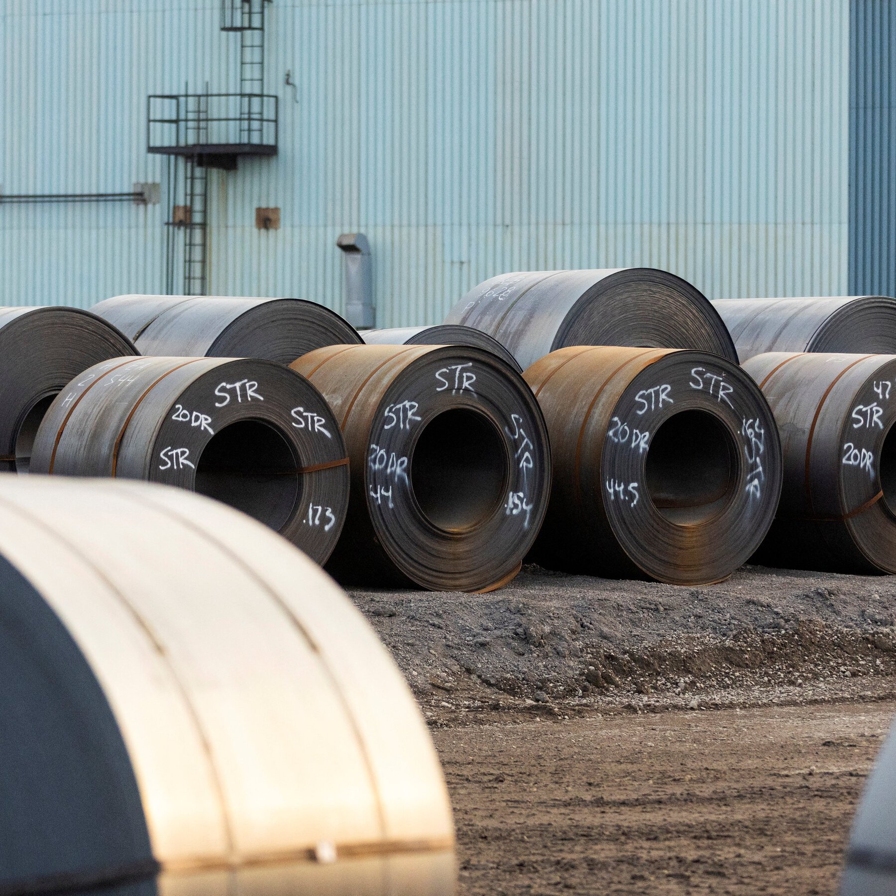 Large steel coils marked with chalk labels sit on a dirt surface outside a light-blue industrial warehouse.