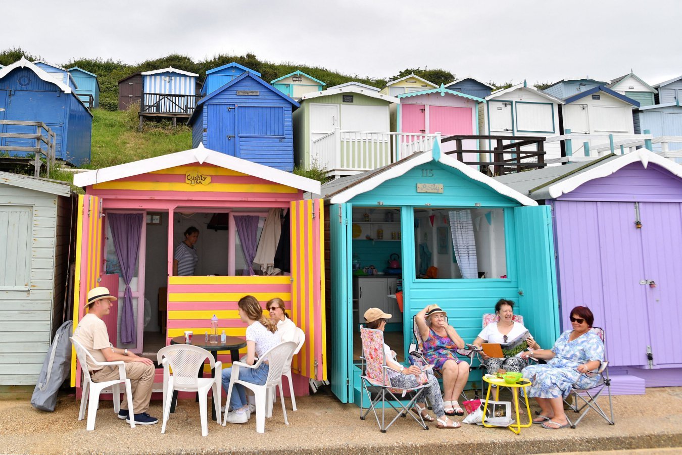 People sit at outdoor tables and chairs in front of colorful wooden beach huts on a hillside, with more huts visible in the background.