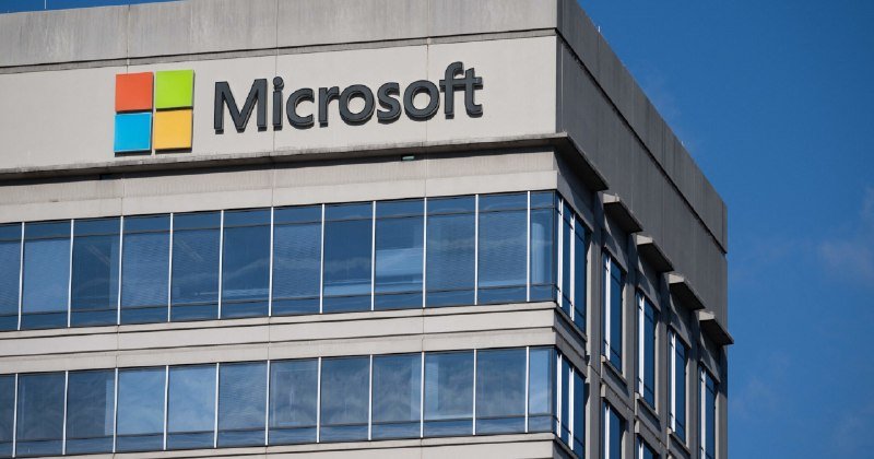 The Microsoft logo with its four-colored square emblem is displayed on the upper corner of a modern building facade featuring tinted glass windows under a clear blue sky.