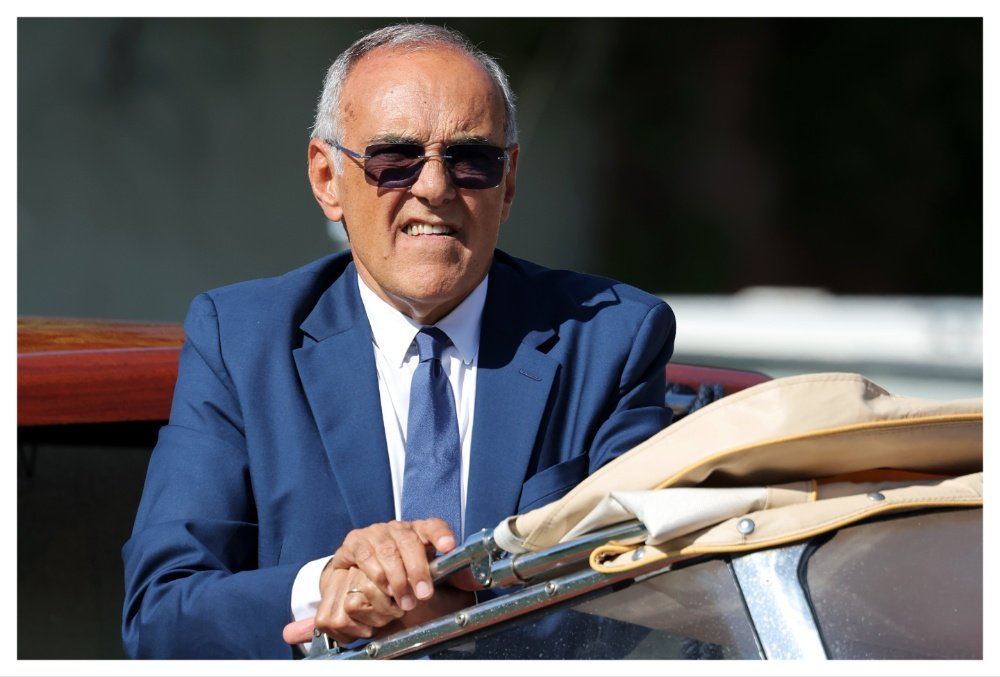 An older man in a blue suit and dark sunglasses smiles while seated in a vehicle with a folded tan convertible top.