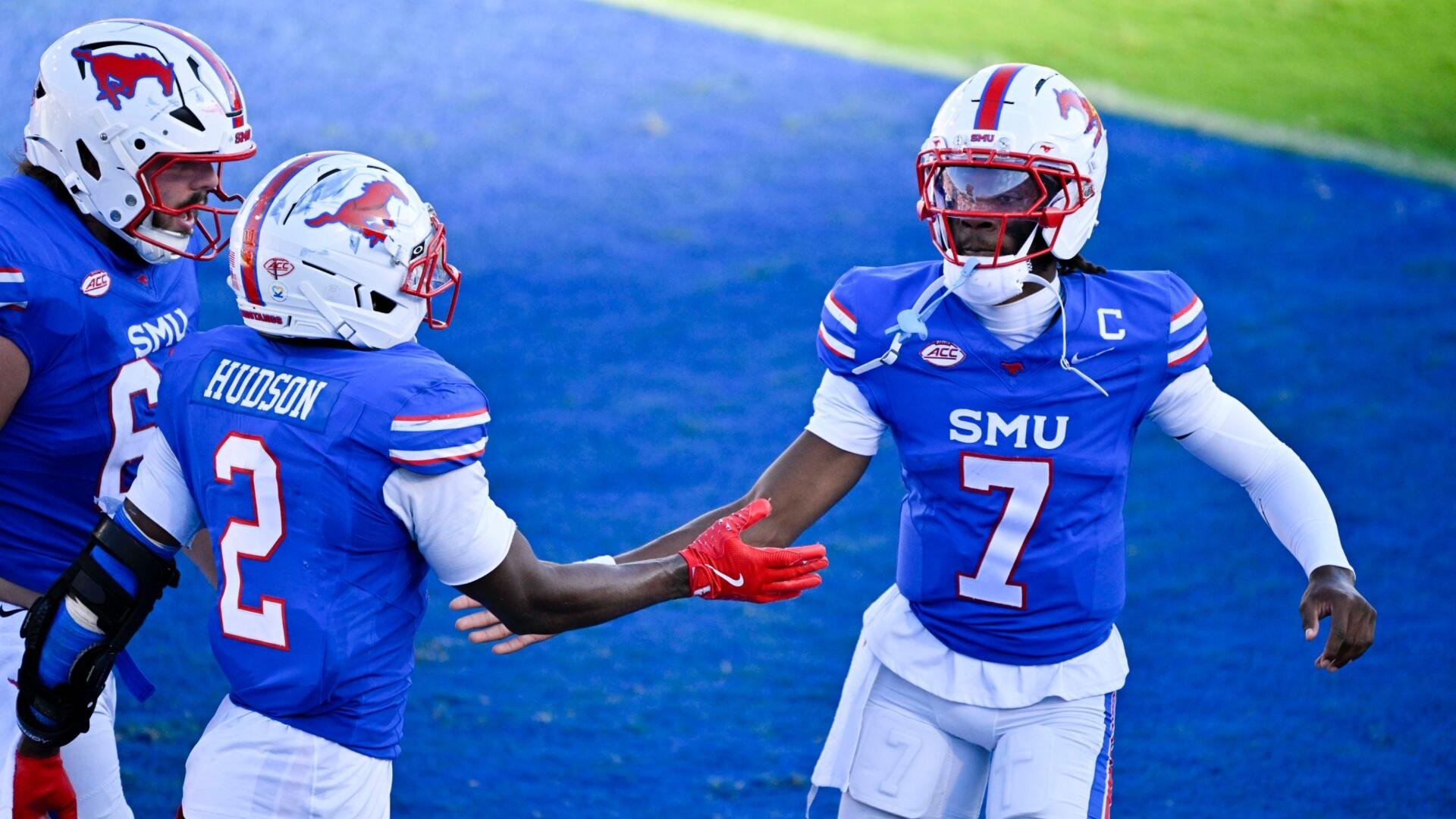 Three SMU football players in blue jerseys and white helmets with a red mustang logo celebrate on a blue turf field, with player #7 exchanging a high-five with a teammate.