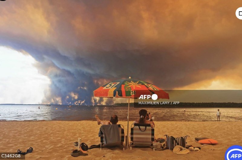Two people sit under a colorful beach umbrella on a sandy shore, photographing a massive plume of smoke rising over the water at sunset.