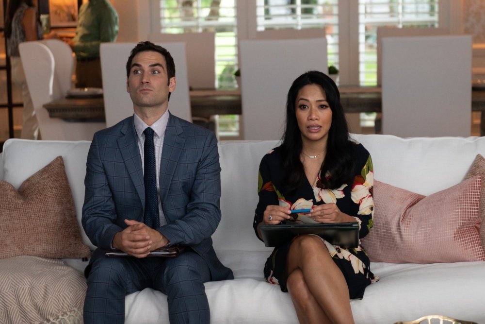 A man in a blue plaid suit and a woman in a floral dress sit on a white couch in a living room, both holding tablets and looking off to the side.