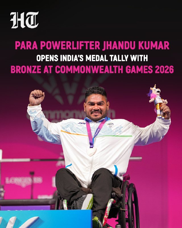 A man in a wheelchair wearing an India team jacket raises his arms while holding a medal and a small mascot figurine on a podium.