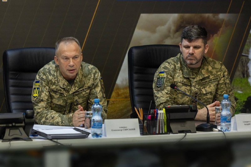 Two men in Ukrainian military camouflage uniforms sit at a conference table with microphones, water bottles, and nameplates, speaking at what appears to be a formal briefing.