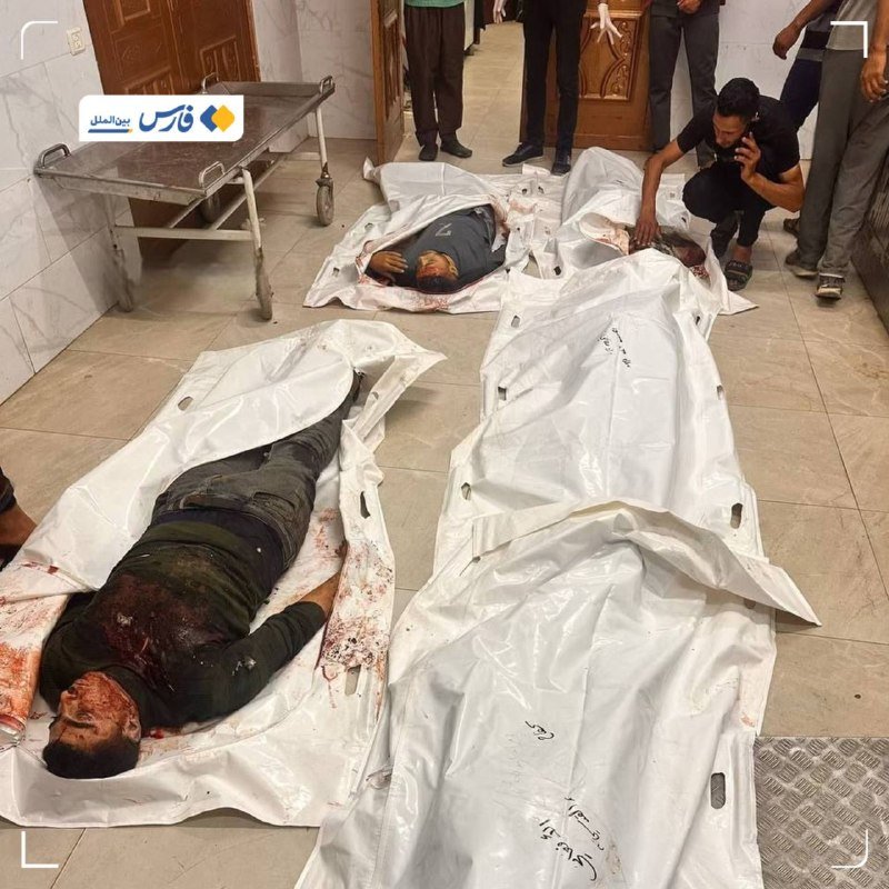 Several bloodied bodies lie in white body bags on a tiled hospital floor, as a man kneels beside them speaking on a phone and others stand nearby.
