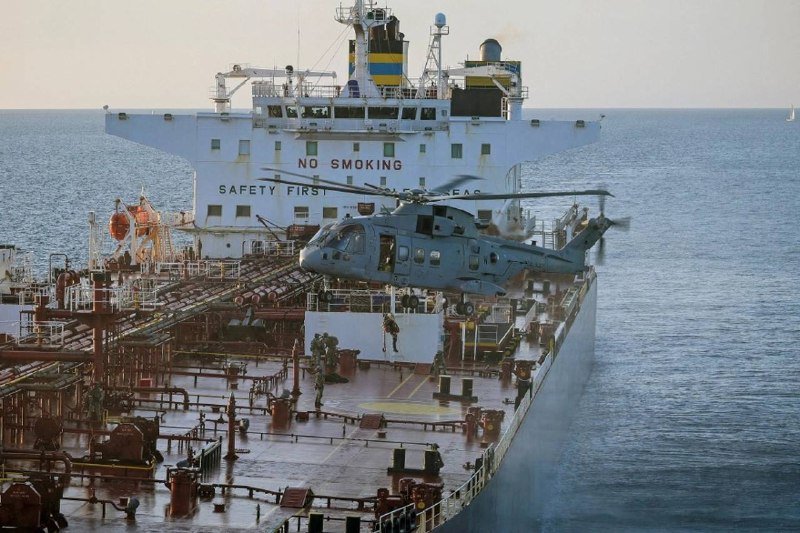 A gray helicopter hovers above the deck of a large cargo ship at sea, with the superstructure reading "NO SMOKING" and "SAFETY FIRST" visible behind it.