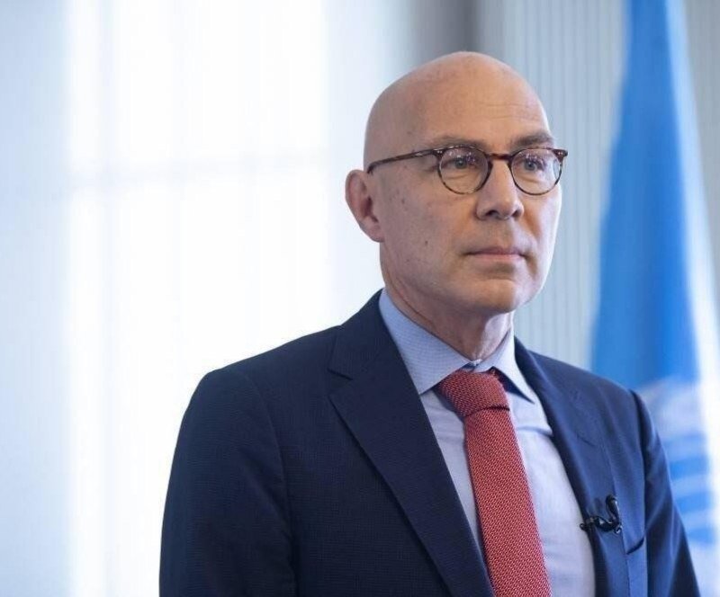 A bald man wearing glasses, a dark blue suit, and a red tie stands in front of a blue flag and a bright window.