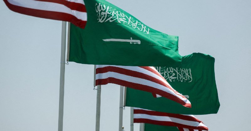 American and Saudi Arabian flags fly side by side on flagpoles against a light sky.