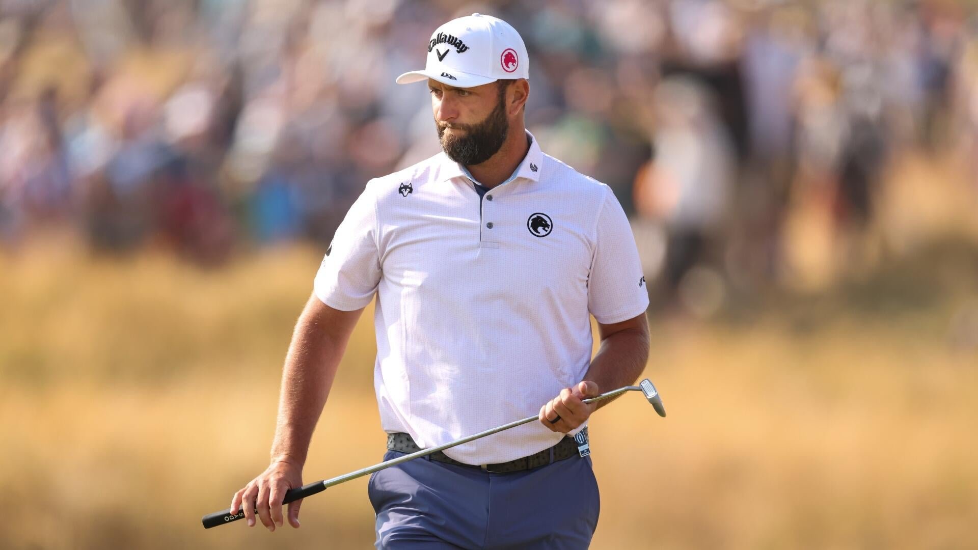 A bearded golfer in a white cap and polo shirt holds a wedge on a grassy course, with blurred spectators visible in the background.