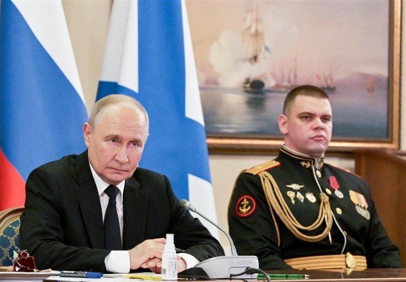 An older man in a dark suit and a younger man in a decorated military dress uniform sit at a table before microphones, with Russian flags and a framed painting of ships behind them.
