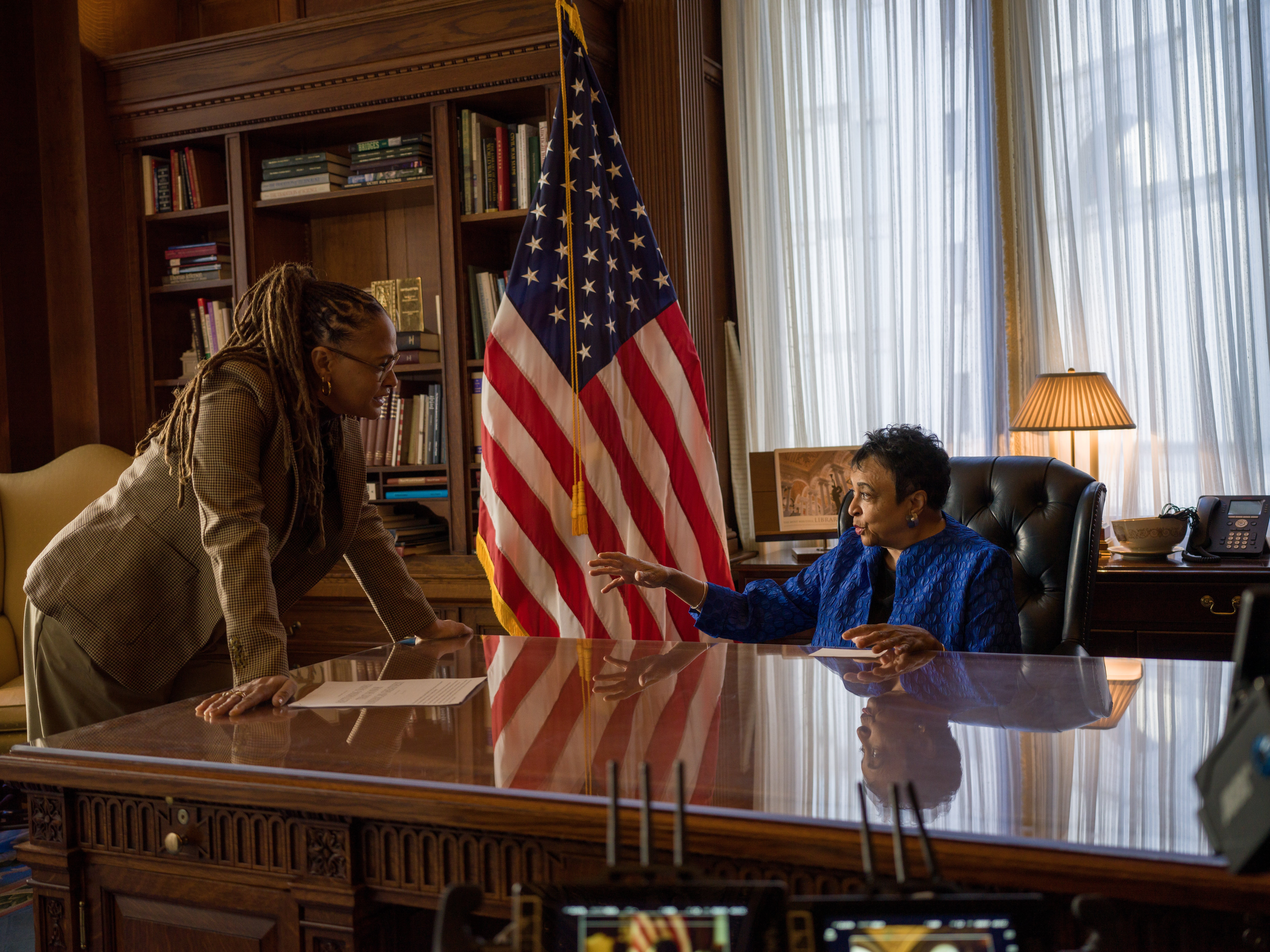Two women converse across a polished wooden desk in a wood-paneled office, with an American flag and bookshelves visible in the background.