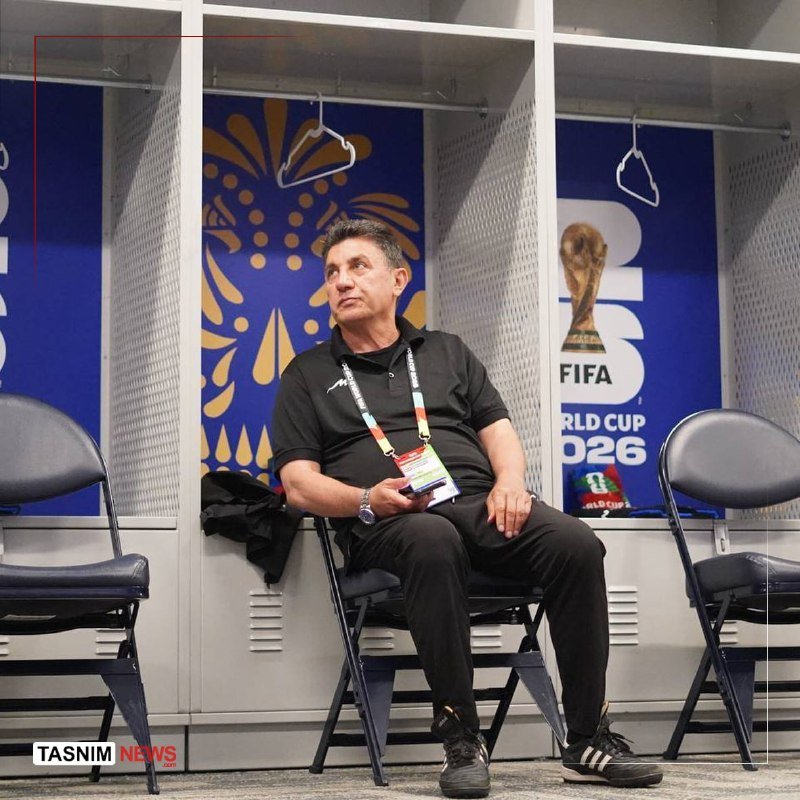 A man wearing a black polo shirt and lanyard sits on a folding chair in a locker room, with FIFA World Cup 2026 branding visible behind him.