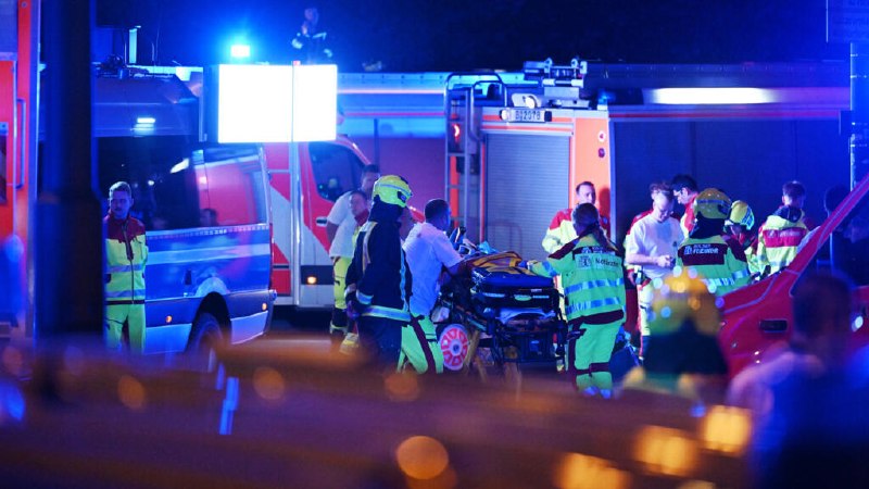 Emergency responders in high-visibility gear surround a stretcher beside a fire truck and ambulance at a nighttime scene illuminated by blue and red lights.
