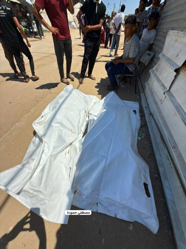 Two body-shaped forms covered in white sheets lie on a dirt ground, surrounded by standing and seated onlookers, with stacked white coffins visible to the right.
