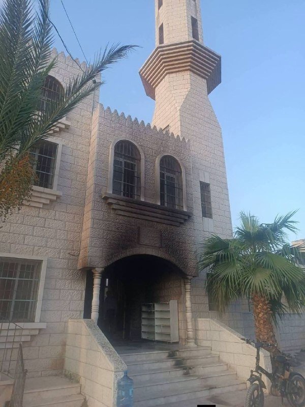 A stone building with a tall minaret, arched windows, and an entrance staircase, flanked by a palm tree and bicycles under a clear blue sky.