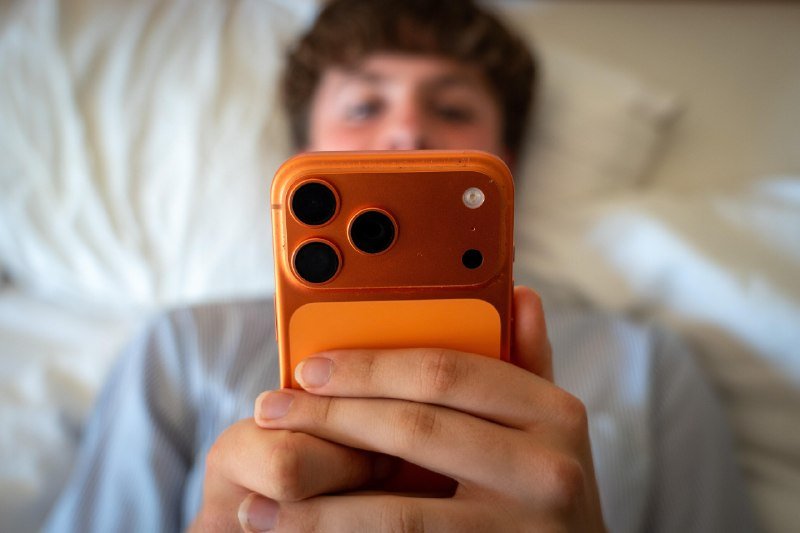 A person holds an orange-cased smartphone with a card slot up to their face while lying in bed.